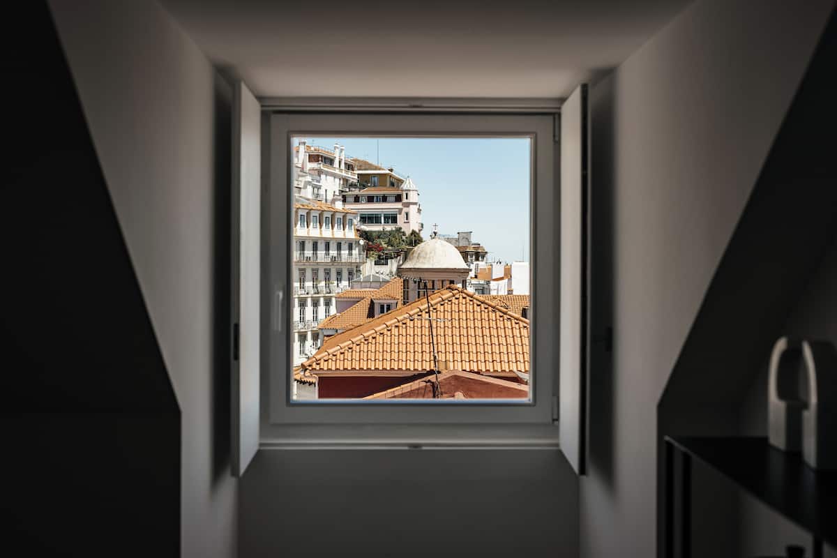 Vista desde una ventana en una habitación con techo inclinado, con vista a un paisaje urbano que incluye edificios y una estructura en forma de cúpula.