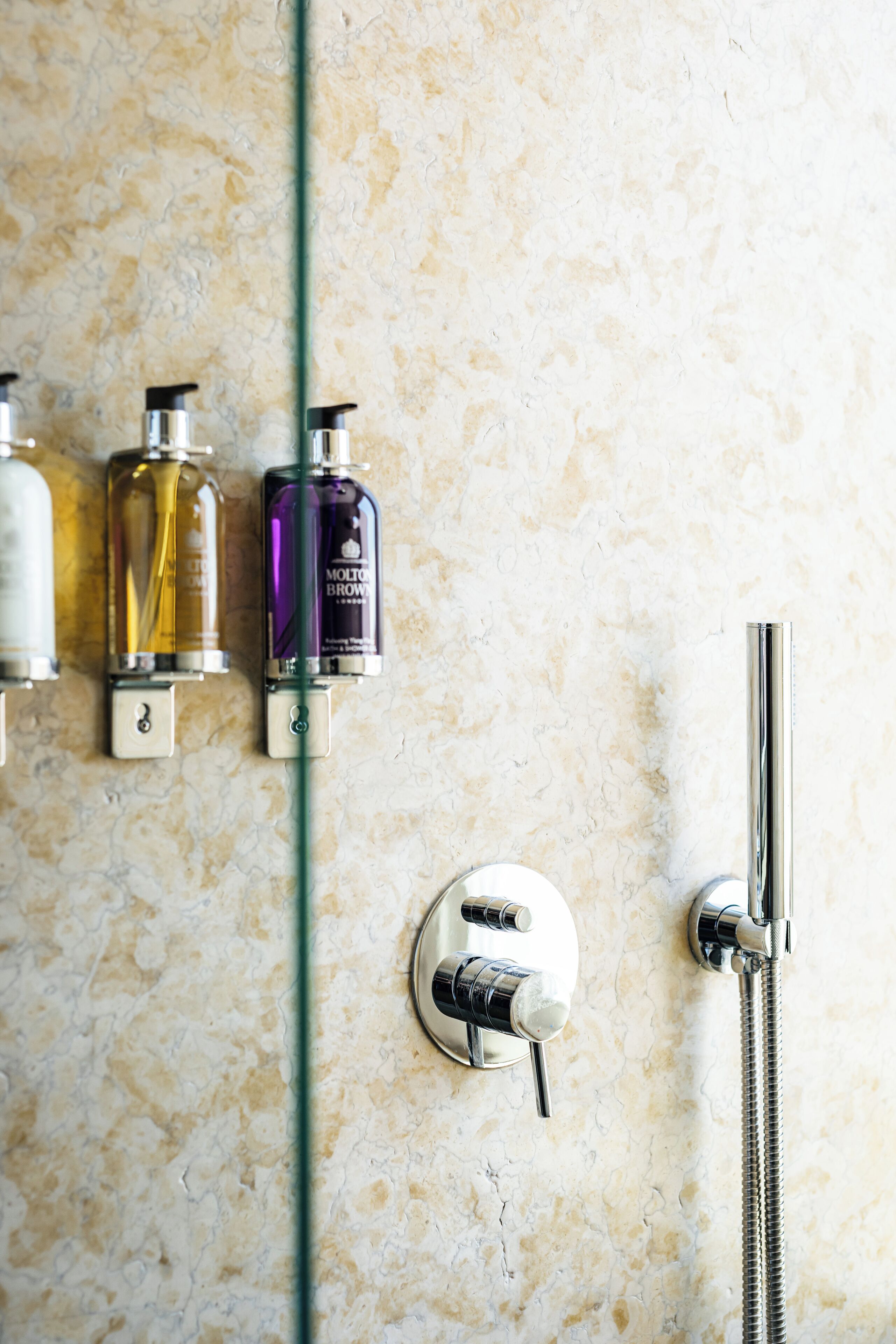A bathroom with a marble wall, a glass shower door, and three bottles of toiletries.