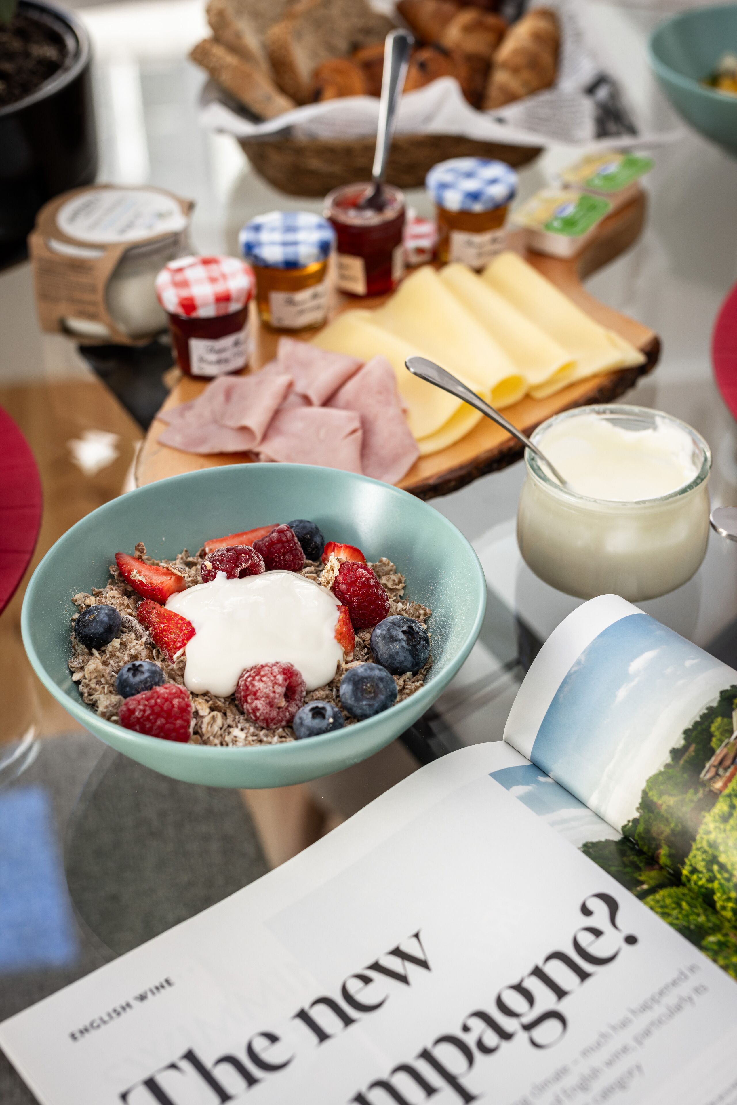 Ein Frühstücksbuffet mit Schüssel Müsli, Joghurt und Beeren, einer Käseplatte mit verschiedenen Käsesorten und Konfitüren, sowie einer Zeitung.