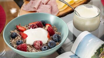 Ein Frühstücksbuffet mit Schüssel Müsli, Joghurt und Beeren, einer Käseplatte mit verschiedenen Käsesorten und Konfitüren, sowie einer Zeitung.