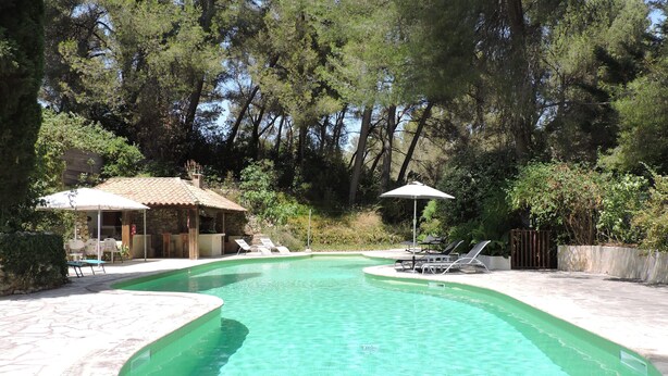 Una piscina con agua cristalina, rodeada de tumbonas y sombrillas, ubicada en un área ajardinada con árboles y un edificio de piedra al fondo.