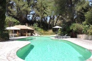 A swimming pool with clear water, surrounded by lounge chairs and umbrellas, set in a landscaped area with trees and a stone building in the background. - Maison de Campagne Avec Piscine Privée (Aubagne)