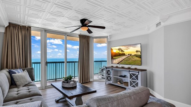 Un salon spacieux doté d’une grande fenêtre donnant sur la mer, d’un ventilateur de plafond et d’un téléviseur posé sur une console en bois.