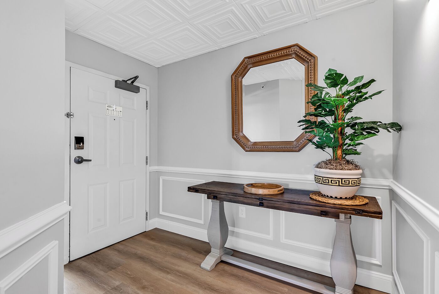 Un couloir avec une porte blanche, une console en bois sur laquelle est posée une plante en pot, un miroir et une petite étagère.