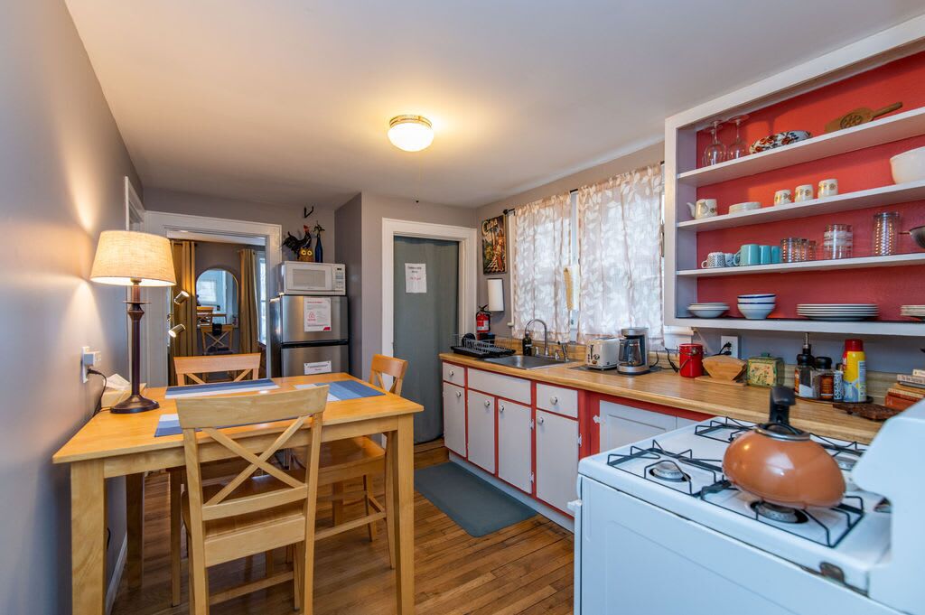 A compact kitchen with a gas stove, wooden dining table, and a red shelf with kitchenware.