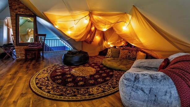 A cozy attic bedroom with a wooden floor, a patterned rug, a plush sofa, and a large mirror.
