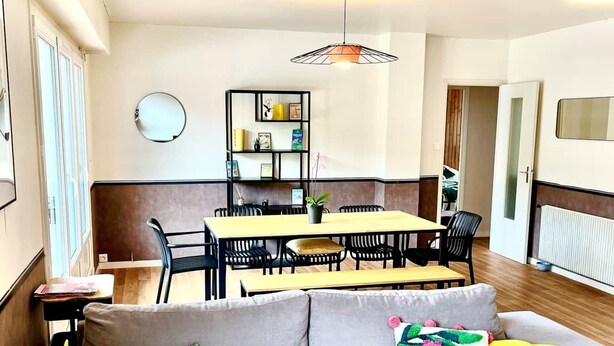 Una sala moderna con un sofá gris, cojines coloridos, una zona de comedor con mesa y sillas, y una lámpara colgante.