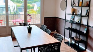 A modern dining area with a wooden table and chairs, a black metal bookshelf, and a view of the outside through large windows.