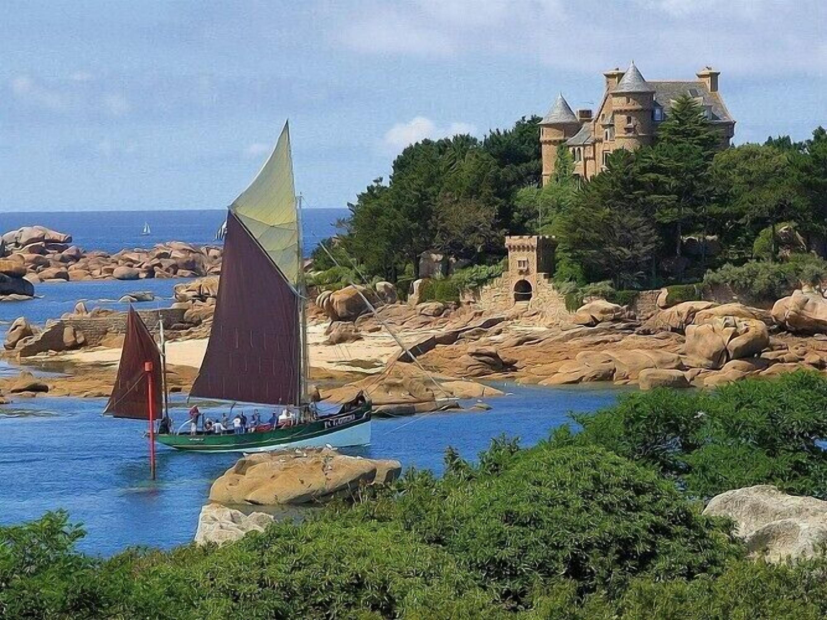 Ein Segelboot mit Passagieren auf ruhigem Gewässer, umgeben von felsigem Gelände und einem schlossähnlichen Gebäude im Hintergrund.