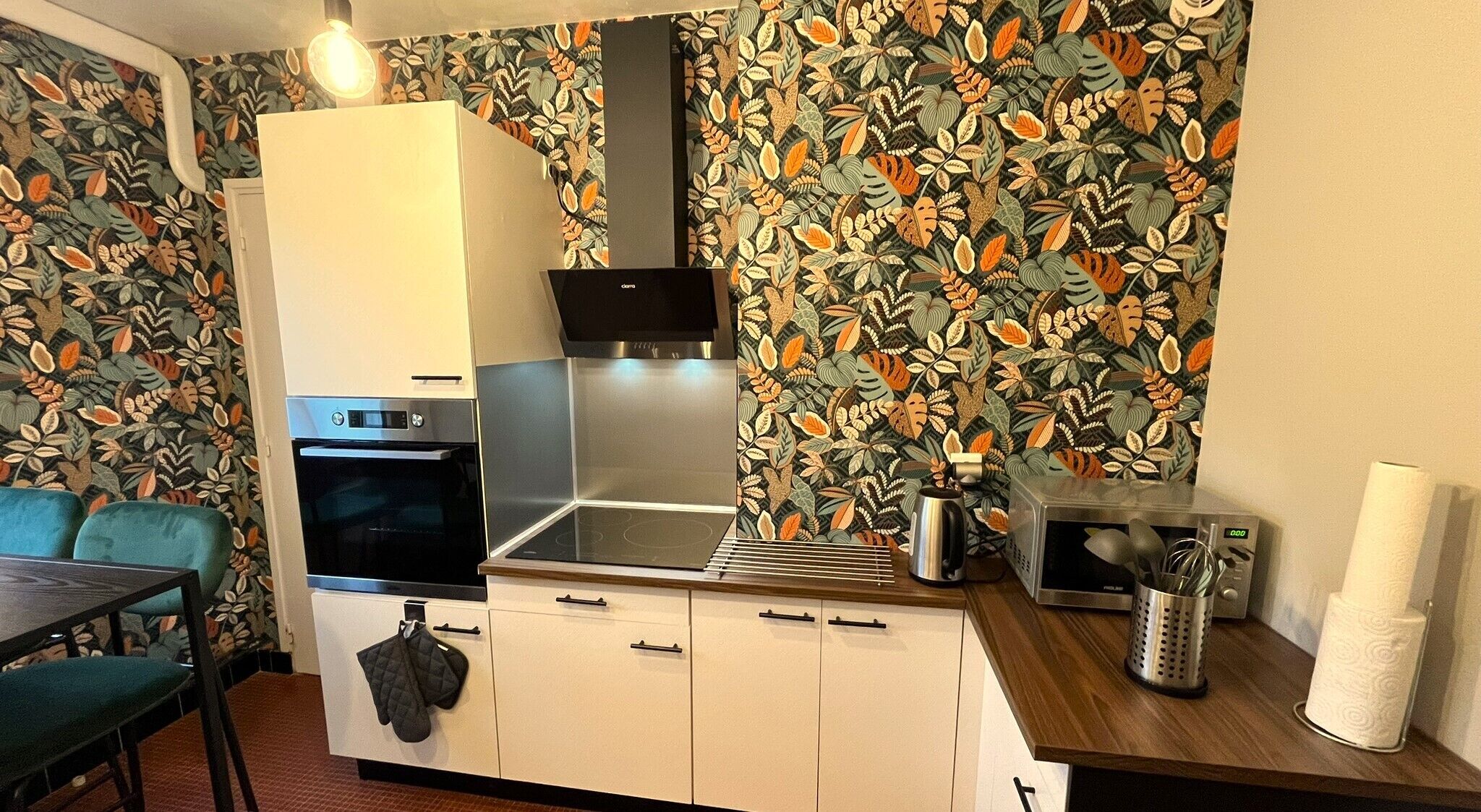 Une cuisine compacte équipée d’un four à micro-ondes, d’un four et d’une bouilloire, avec un papier peint à motifs et un plan de travail en bois.
