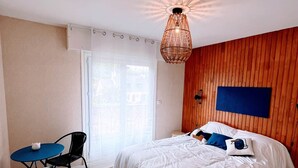 A bedroom with a large bed, a desk with chairs, a hanging light fixture, and a wooden wall panel.