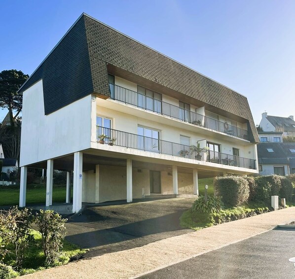 A two-story building with a flat roof, a balcony on the upper floor, and a covered parking area.