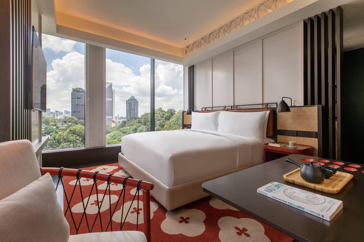 A modern hotel room with a large bed, a desk with a teapot, a view of the city, and a red patterned rug.