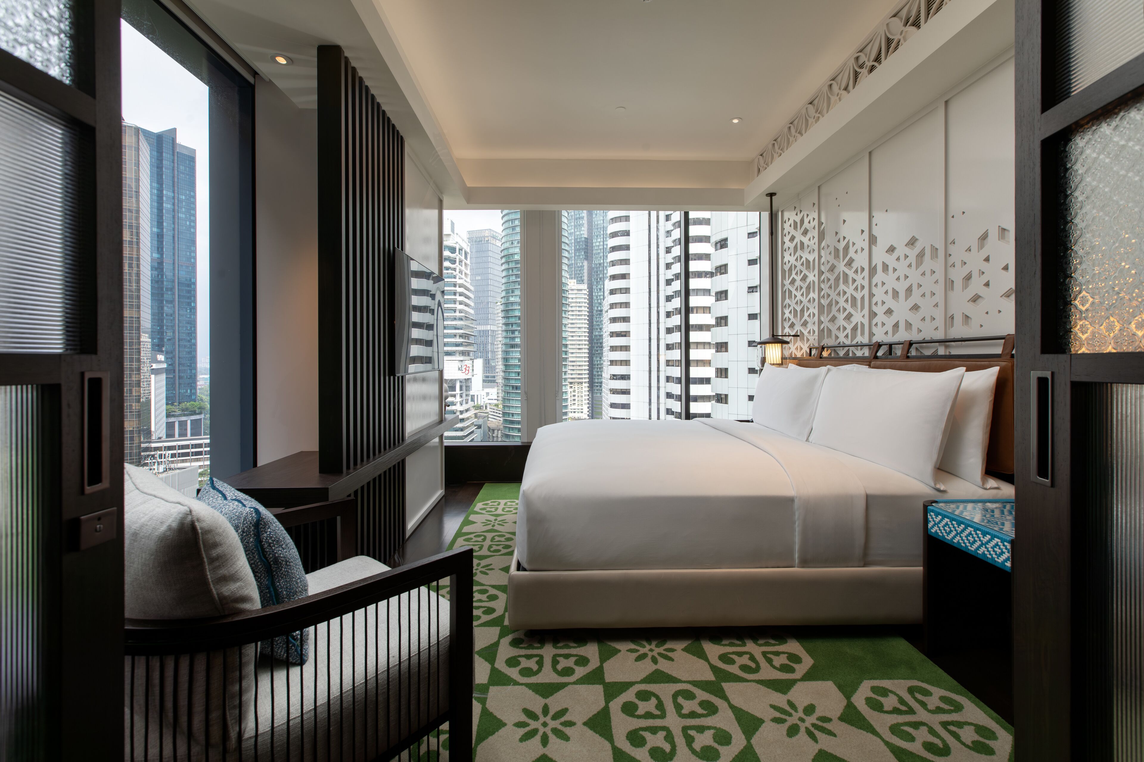 Una moderna camera d'hotel con un letto grande, vista su alti edifici e un tappeto verde con motivo.