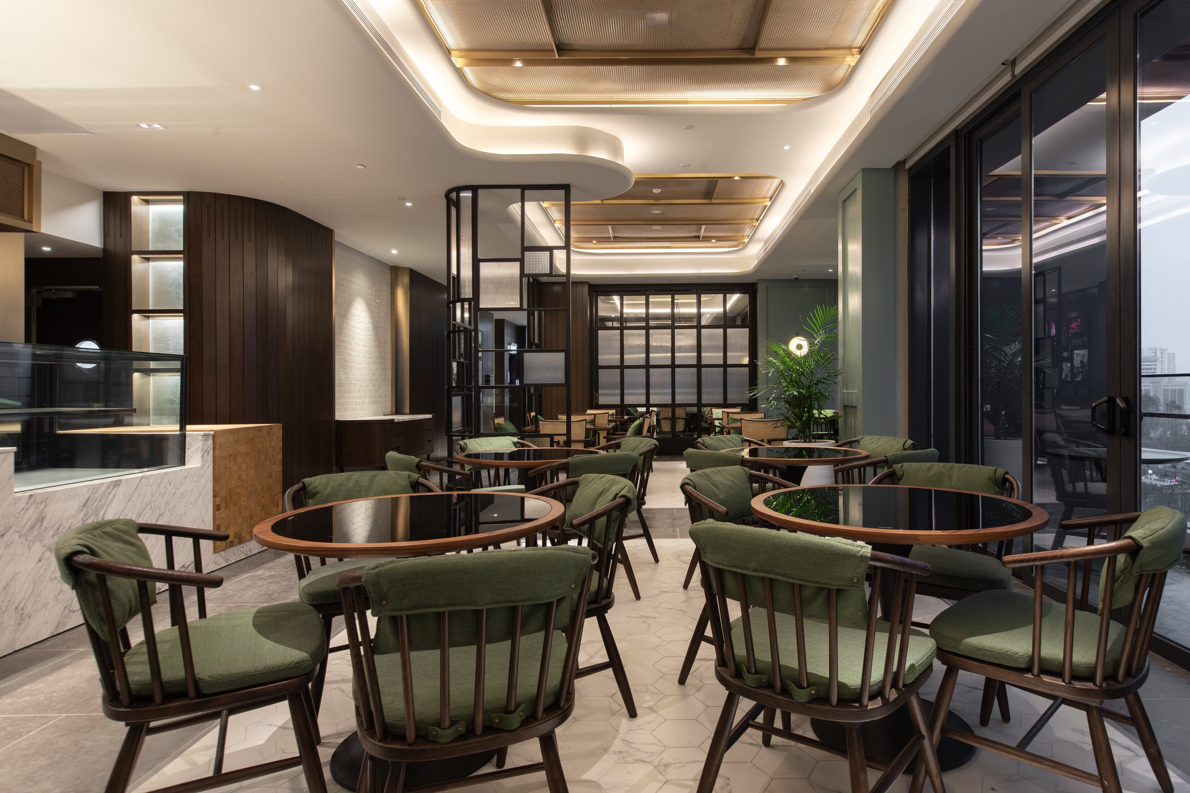 A modern interior with round tables and green upholstered chairs, a bar area, and large windows offering a view.