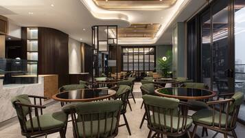 A modern interior with round tables and green upholstered chairs, a bar area, and large windows offering a view.