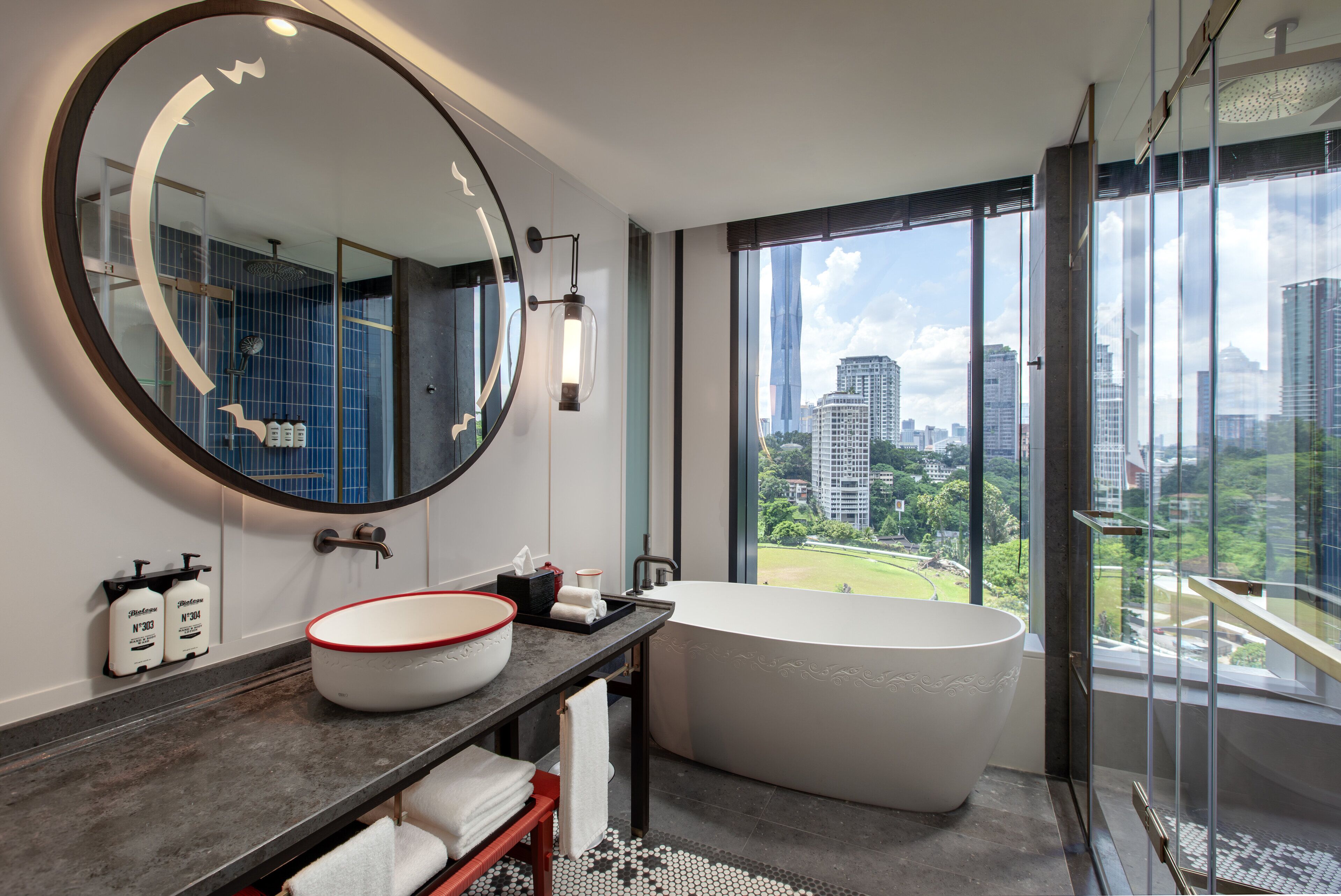 A modern bathroom with a large round mirror, a freestanding bathtub, and a large window offering a city view.