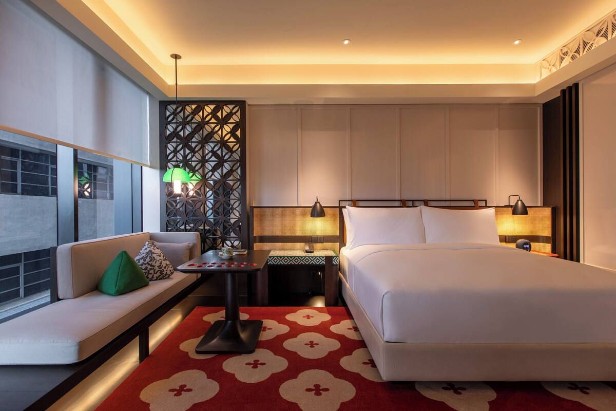 A modern hotel room with a large bed, a seating area with a sofa and a small table, a decorative partition, and a view of the city.