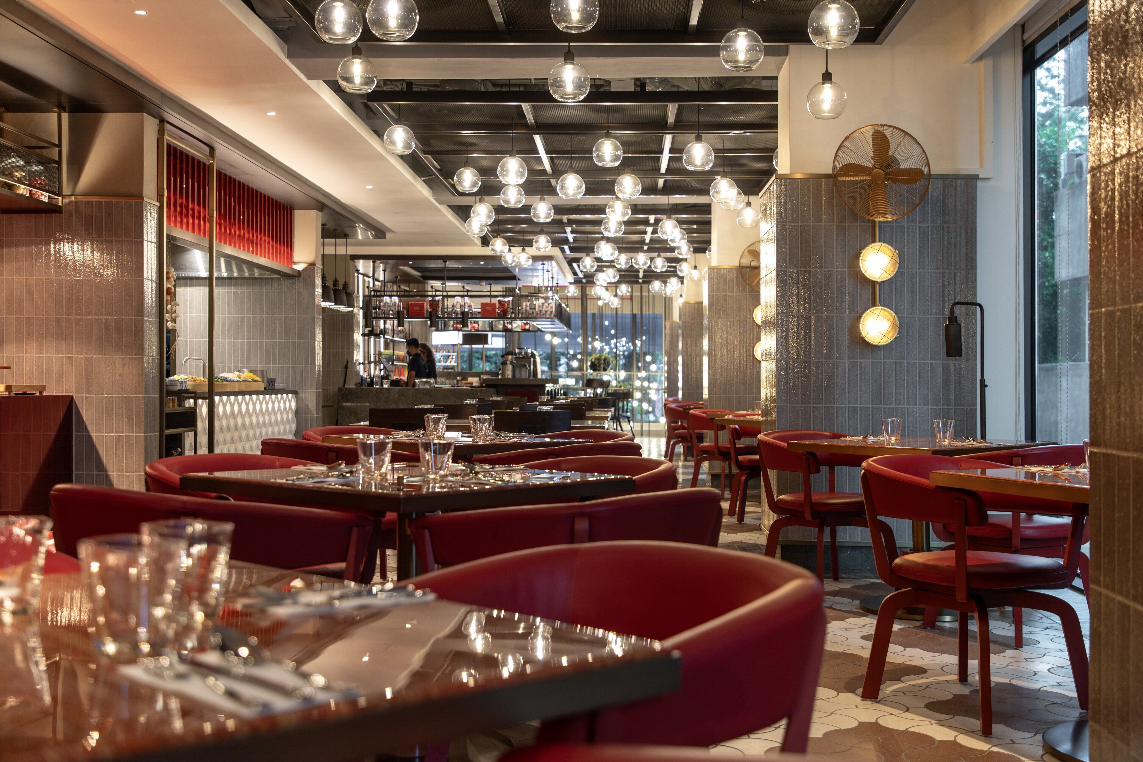 A modern restaurant interior with multiple dining tables and red chairs, overhead spherical light fixtures, and a visible kitchen area in the background.