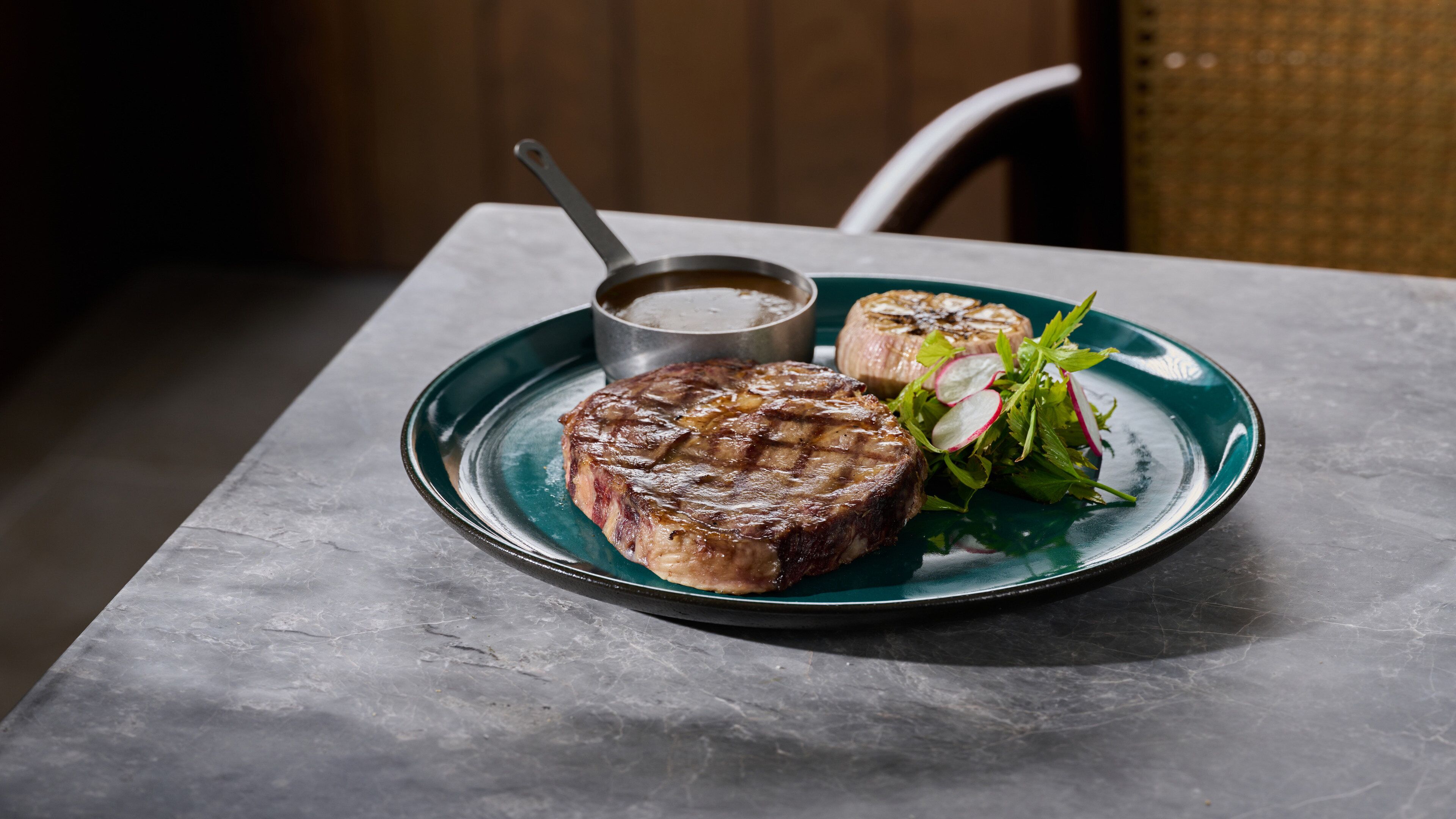 A plate with a grilled steak, a small bowl of sauce, and a side salad on a marble table.