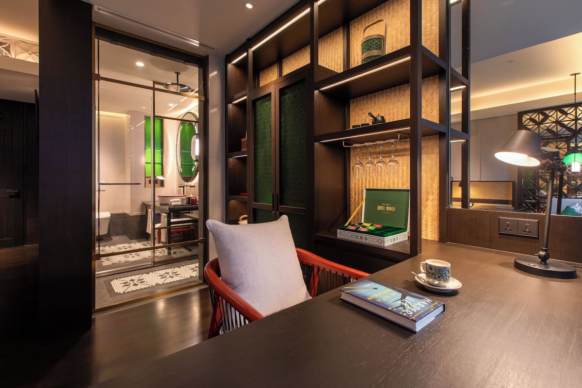 A modern hotel room with a wooden desk, a comfortable chair, a bookshelf, and a large window offering a view of the interior.