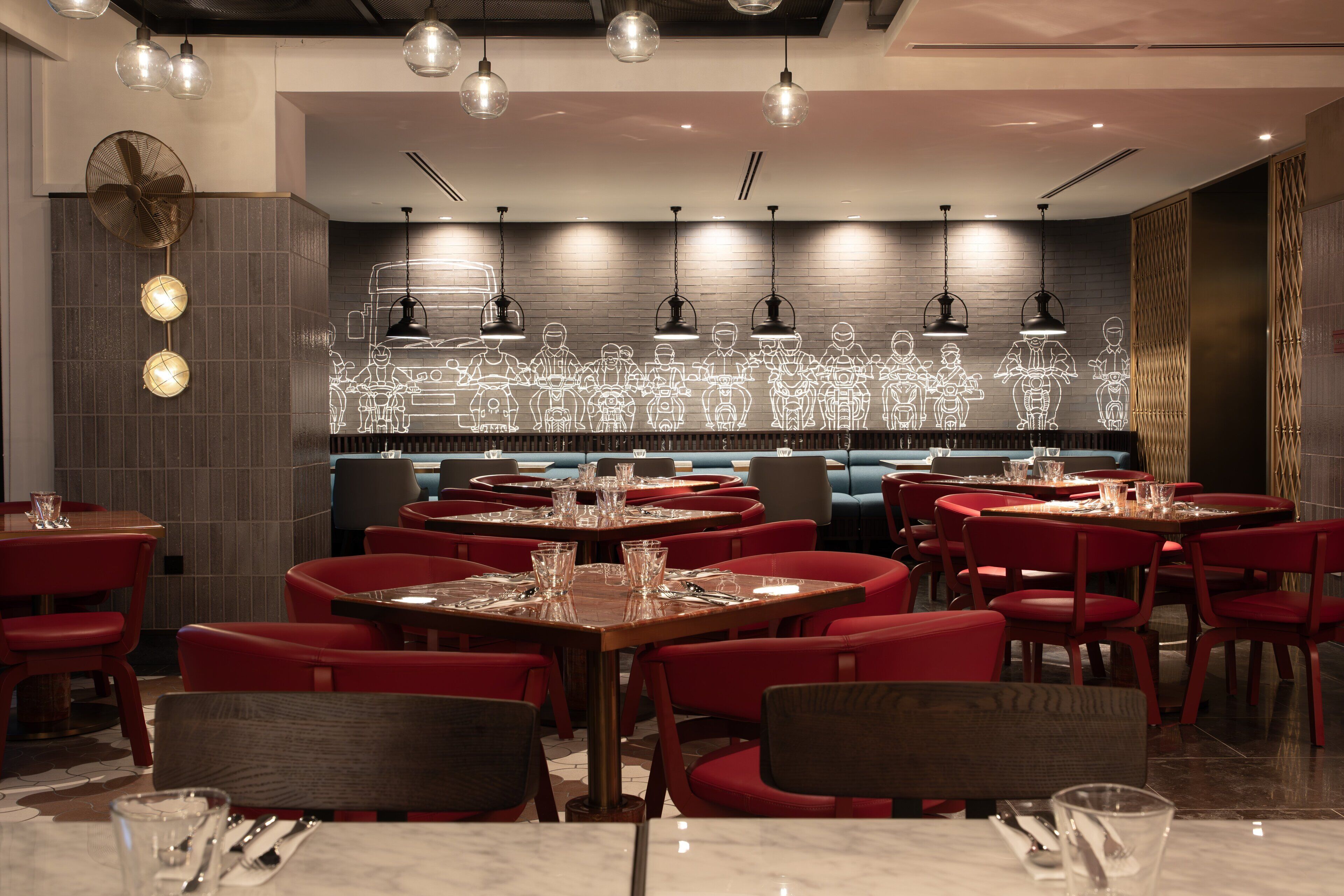 A restaurant with multiple tables and red chairs, a patterned wall, and hanging pendant lights.