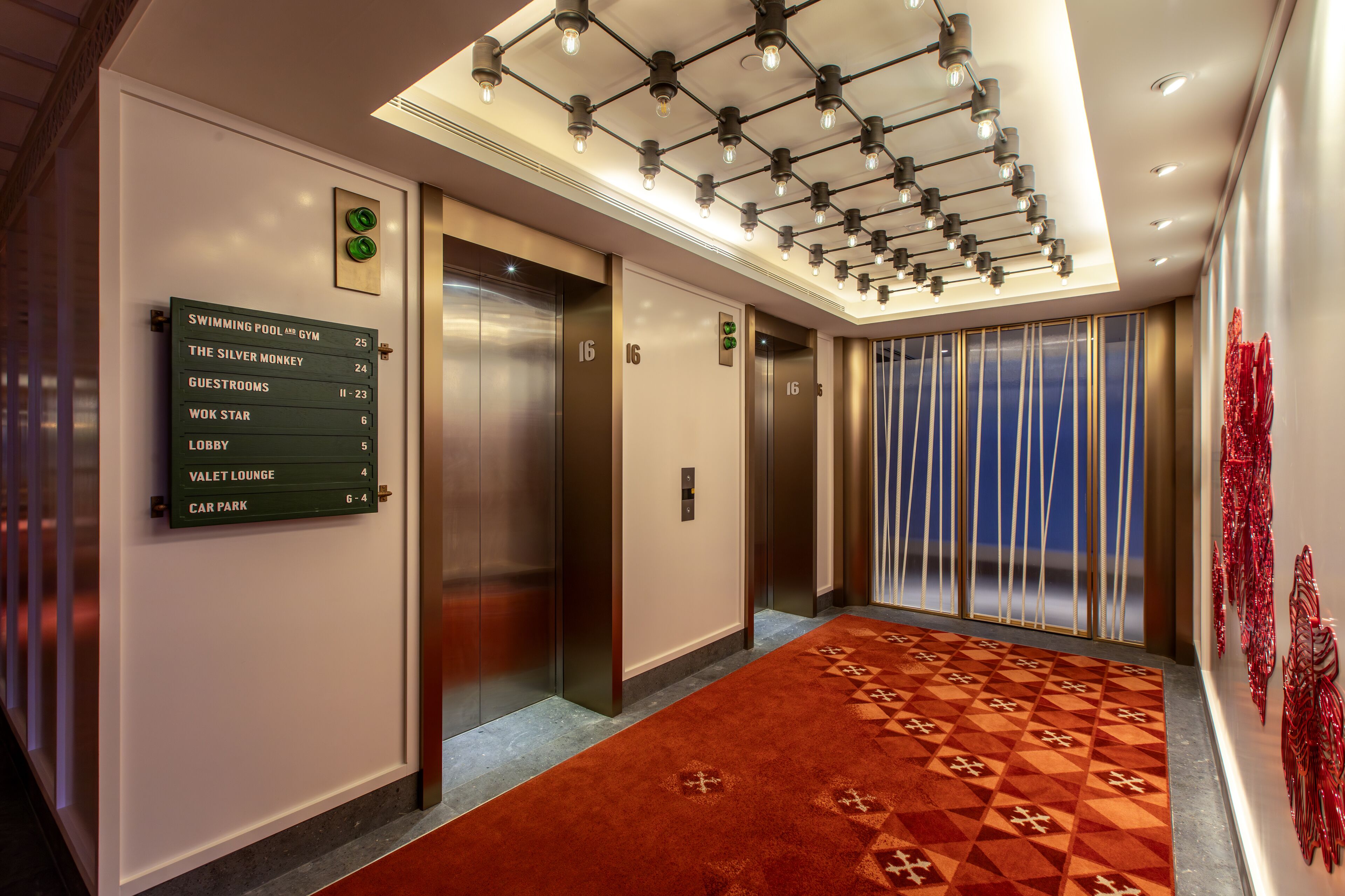 A hotel lobby with a red patterned carpet, a green sign listing amenities, and three elevators.