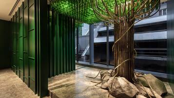 A modern interior with a unique tree sculpture made of green bottles, a staircase with green doors, and a view of a building outside.