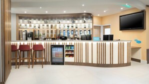 A hotel reception area with a counter, bar stools, a beverage dispenser, and a television mounted on the wall. - Hampton by Hilton London Old Street (London)