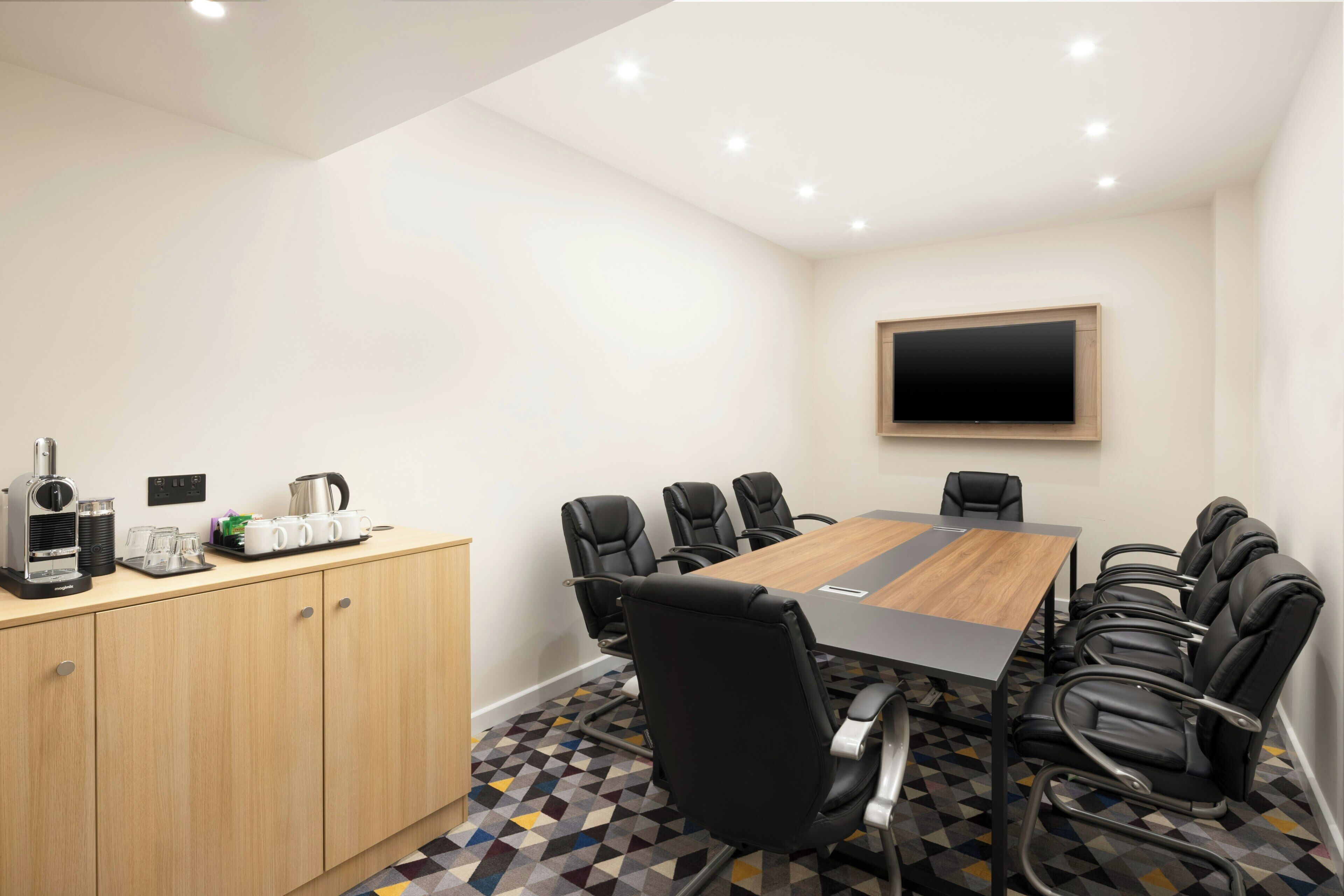 Une salle de conférence avec une longue table, des chaises noires, un coin café et un téléviseur fixé au mur.