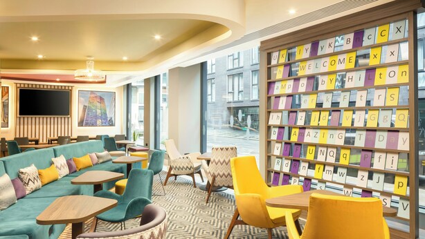 A modern lounge area with a sofa, chairs, tables, a bookshelf with alphabet letters, and a TV screen. - Hampton by Hilton London Old Street (London)