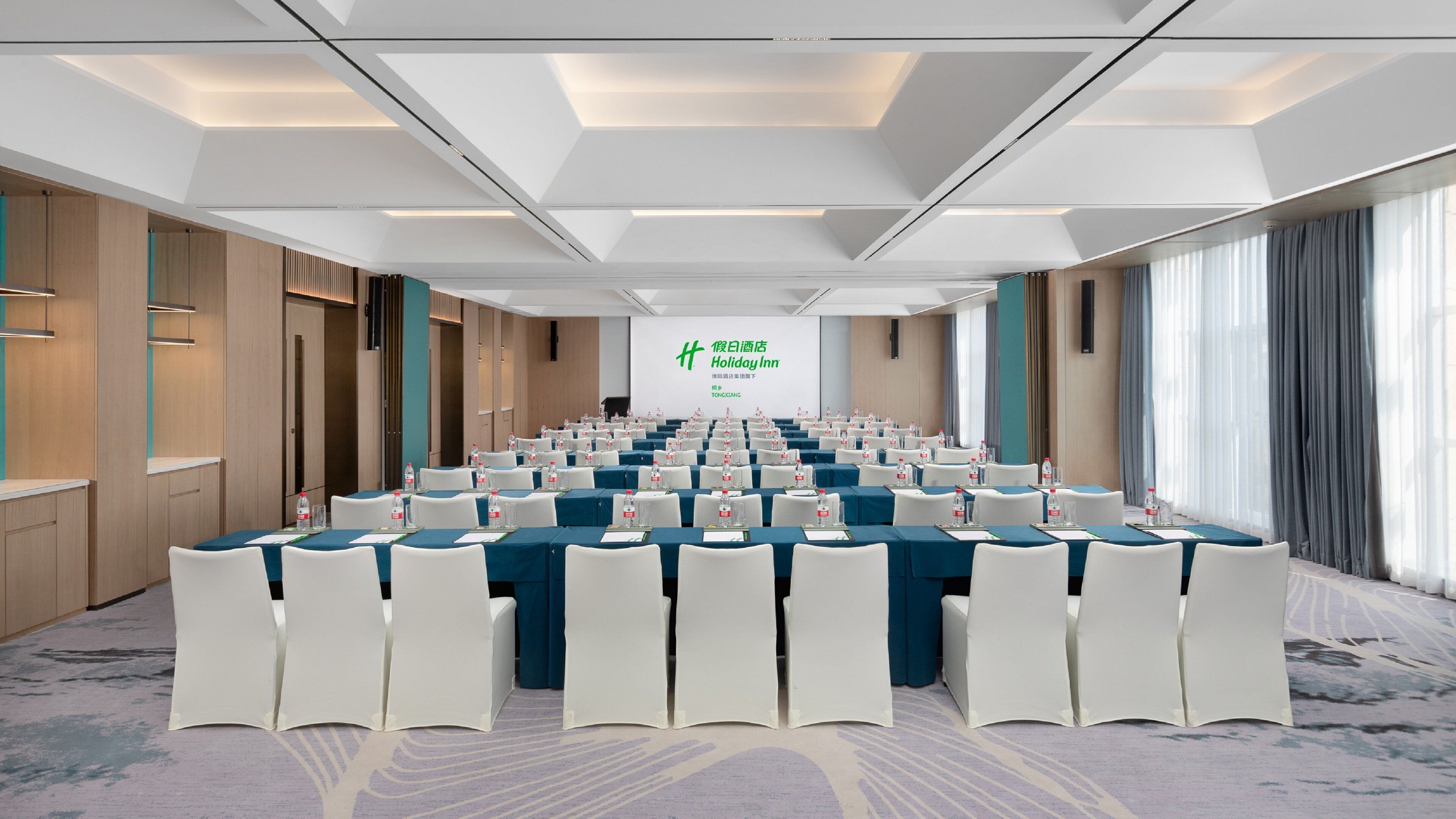 A conference room with rows of tables and chairs, a projection screen at the front, and a large window on one side.