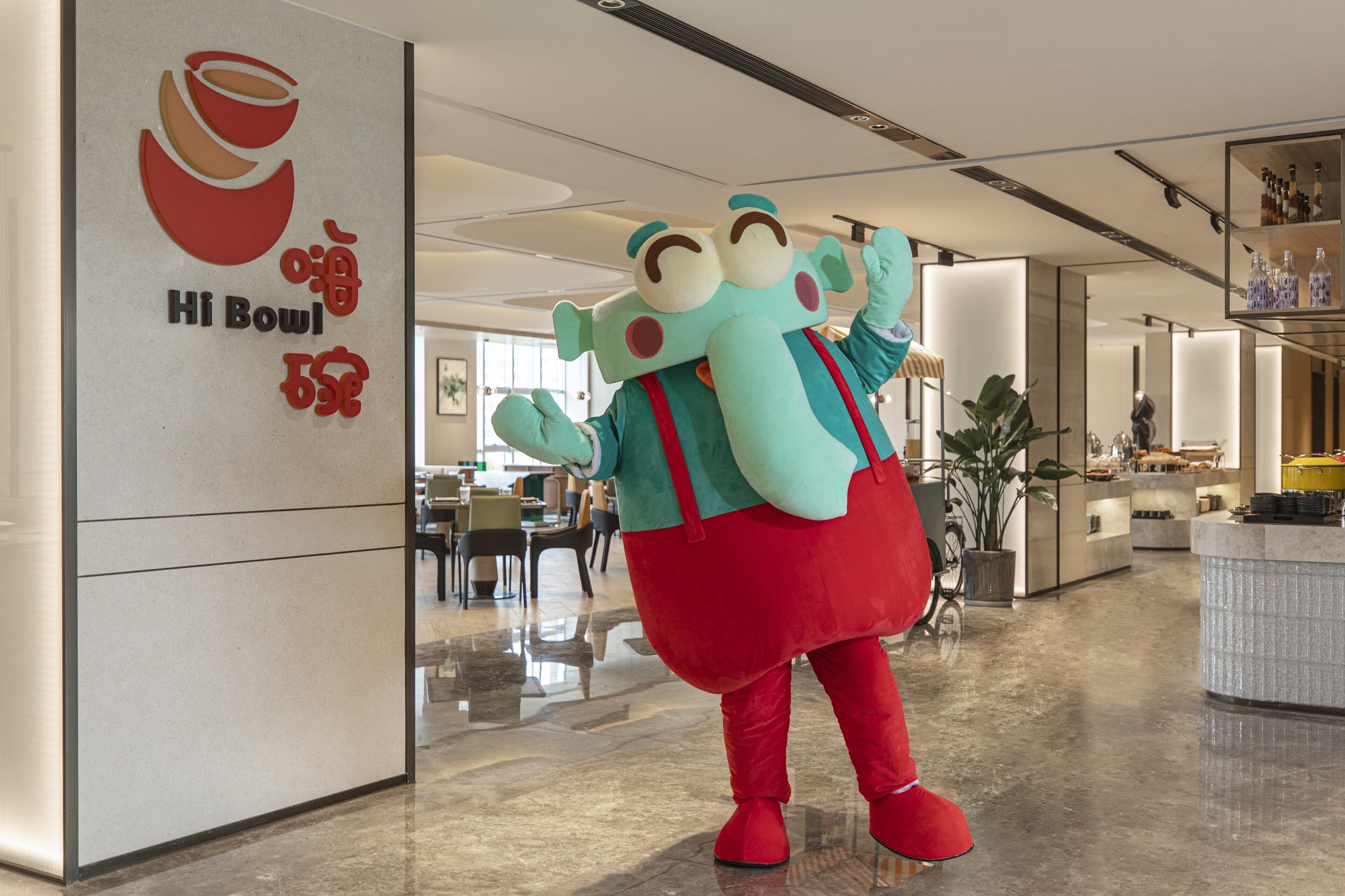 Um personagem de mascote em traje vermelho e azul-turquesa está em um saguão moderno de hotel, com piso lustroso e uma grande planta em vaso.