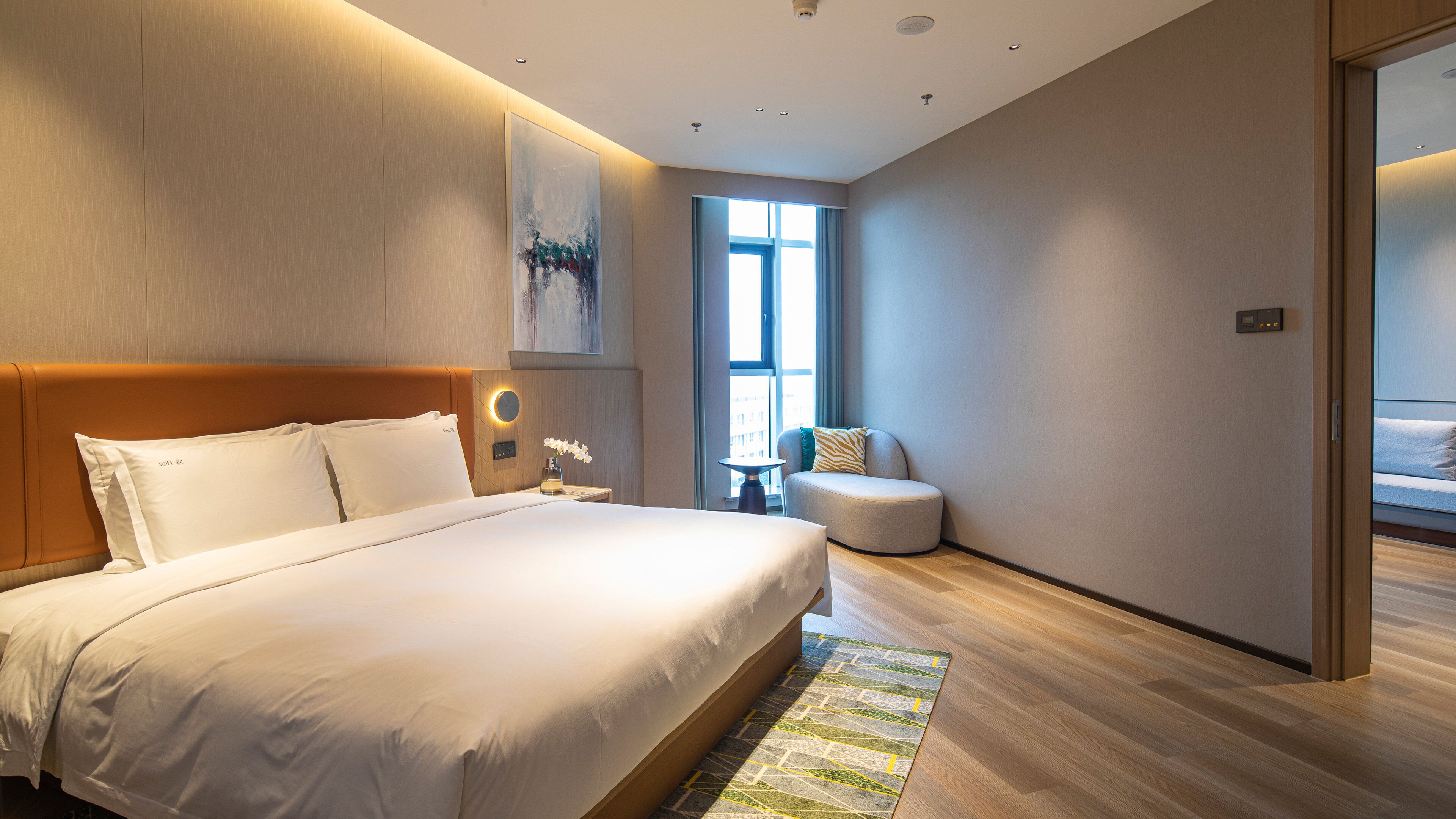 Et moderne hotelværelse med en stor seng, et siddeområde med en stol og et lille bord, samt et stort vindue der giver naturligt lys.