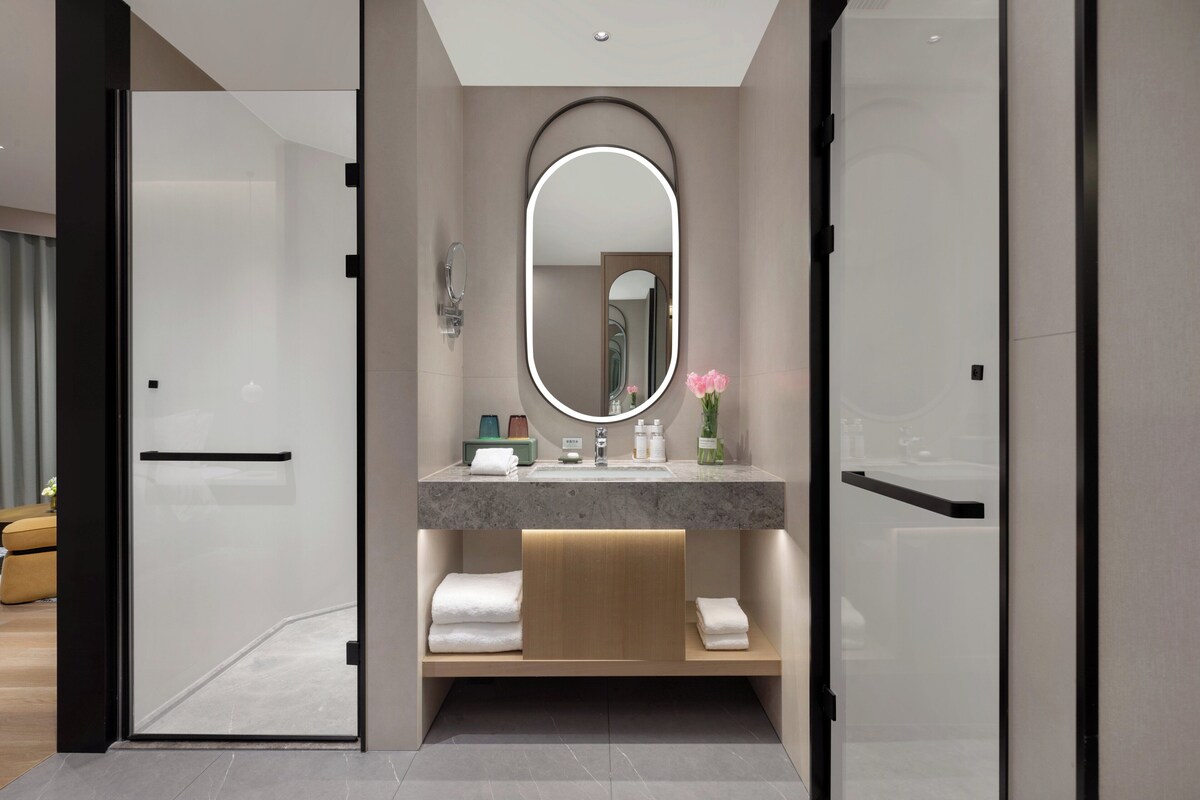 A modern bathroom with a glass door, a round mirror, a marble countertop, and a wooden shelf with towels.