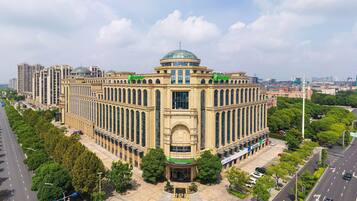 A large, historic building with a central dome and multiple wings, surrounded by trees and modern high-rise buildings.