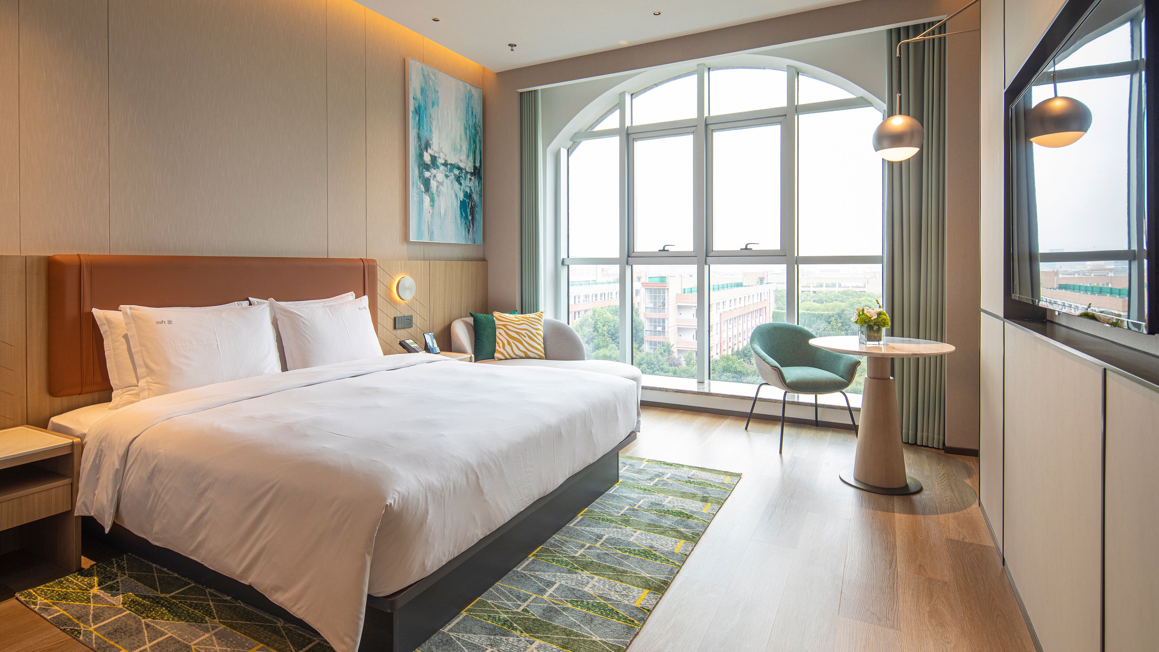Una camera d'albergo moderna con un letto grande, un angolo salotto con una sedia e un tavolo, e un'ampia finestra che offre vista sulla città.