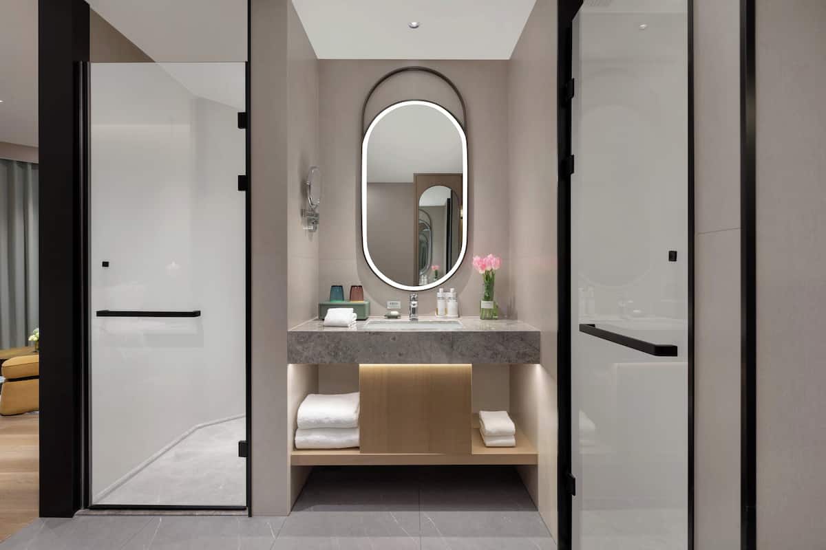 Un baño moderno con puerta de cristal, espejo redondo, encimera de mármol y estante de madera con toallas.