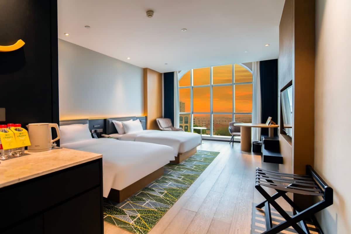 Habitación de hotel con dos camas, un escritorio, una silla, un ventanal amplio con vistas a la ciudad y una mesita.