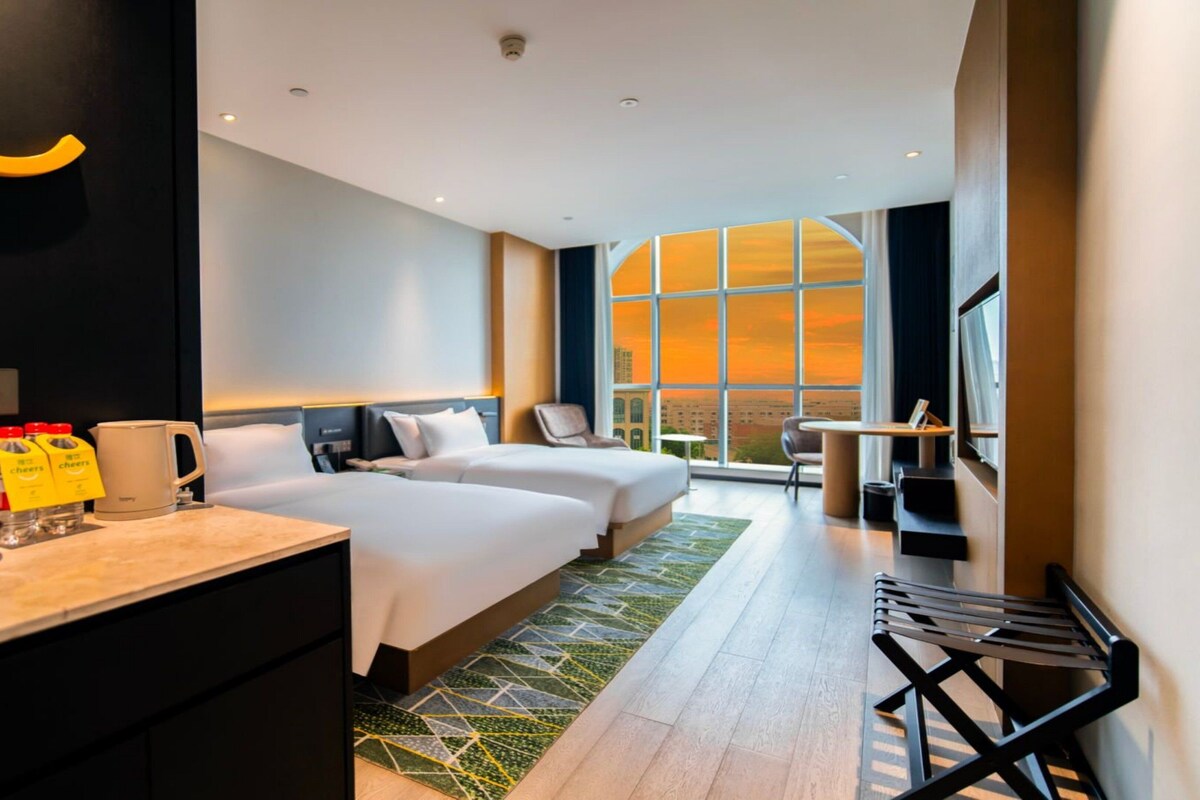 A hotel room with two beds, a desk, a chair, a large window with a city view, and a small table.