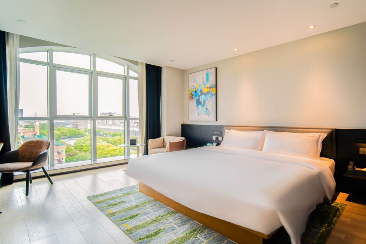 A modern hotel room with a large bed, a desk, and a chair. There is a large window offering a city view.