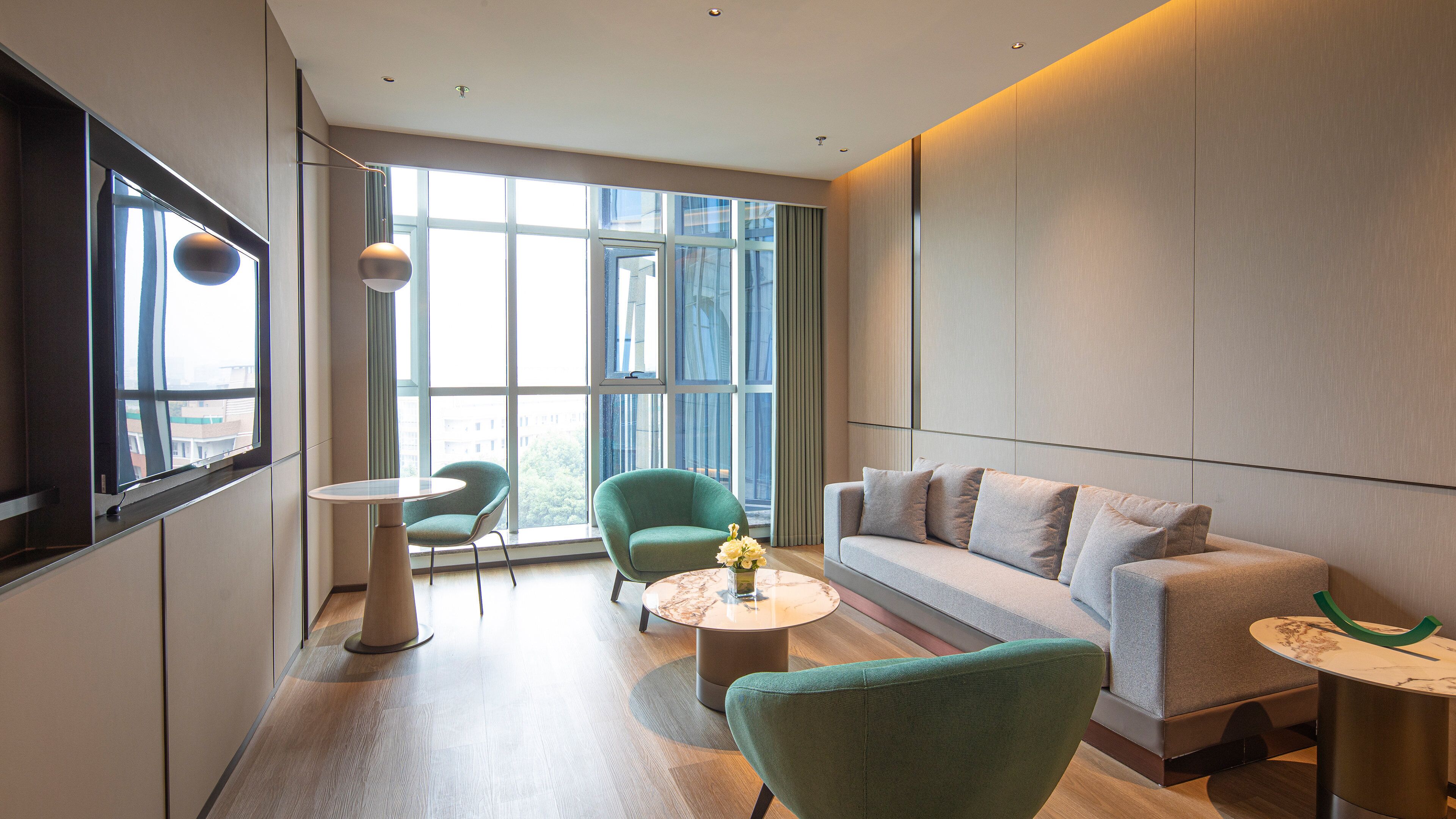 Een moderne hotelkamer met een groot raam, een groene fauteuil, een grijze bank en een klein rond tafeltje met een bloevas.