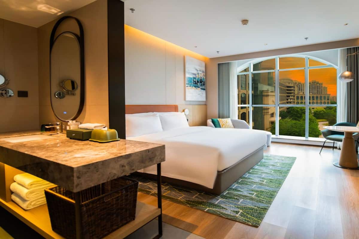 Una habitación de hotel moderna con una cama grande, una zona de estar con una mesa, un ventanal con vistas a la ciudad y un baño con encimera de mármol.