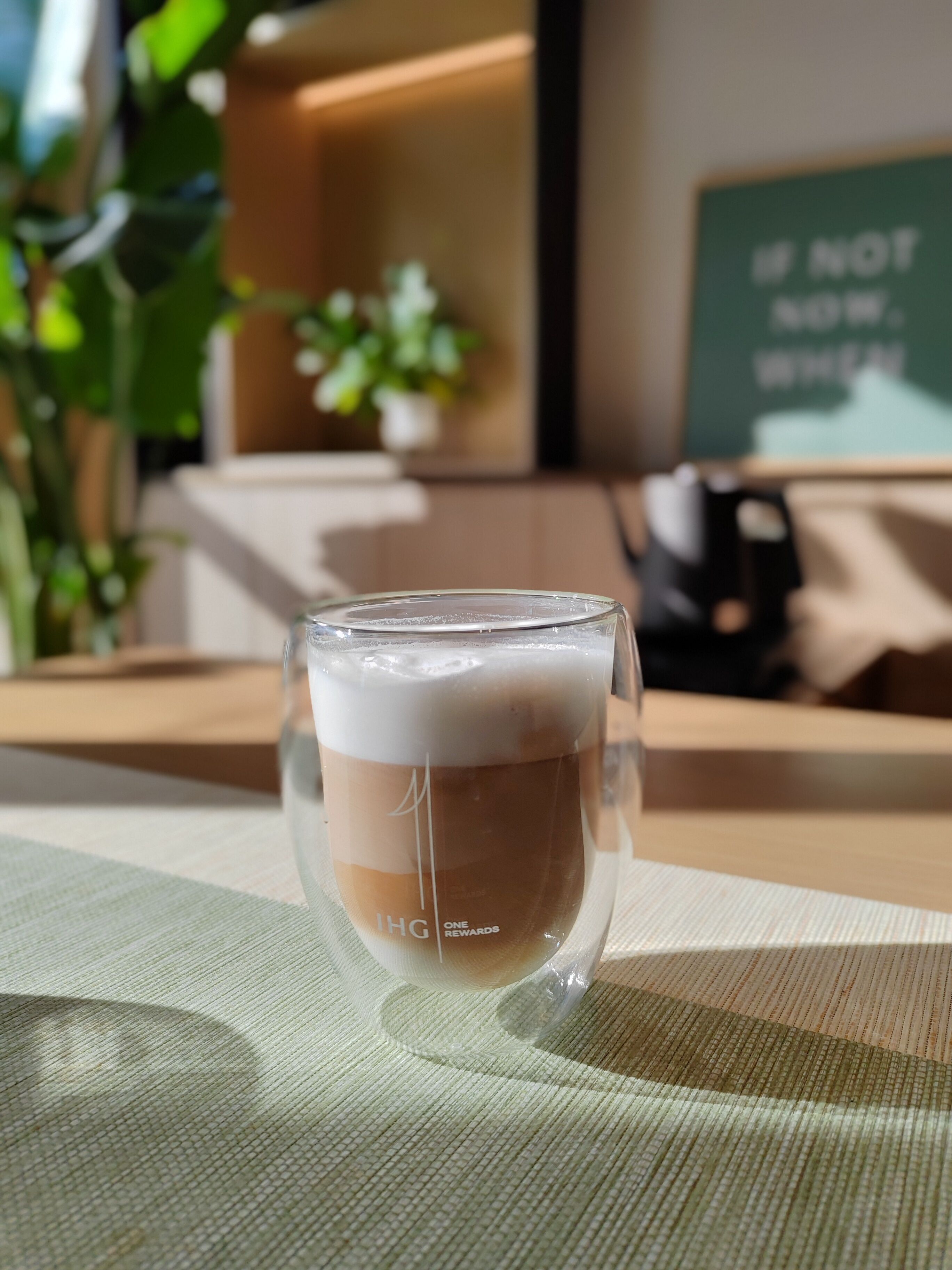 A glass of iced coffee on a table with a placemat, a potted plant, and a sign in the background.