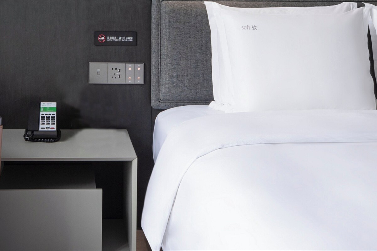 A hotel room with a bed, a bedside table with a phone, and a wall-mounted control panel.