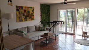 Un salon comprenant un canapé, une table basse et un coin repas avec une table et des chaises. Une grande fenêtre offre une vue sur la verdure environnante.