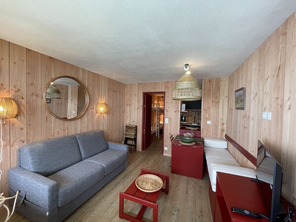 A room with a grey sofa, a red coffee table, a red sofa, a TV, and a kitchen area visible through an open door.