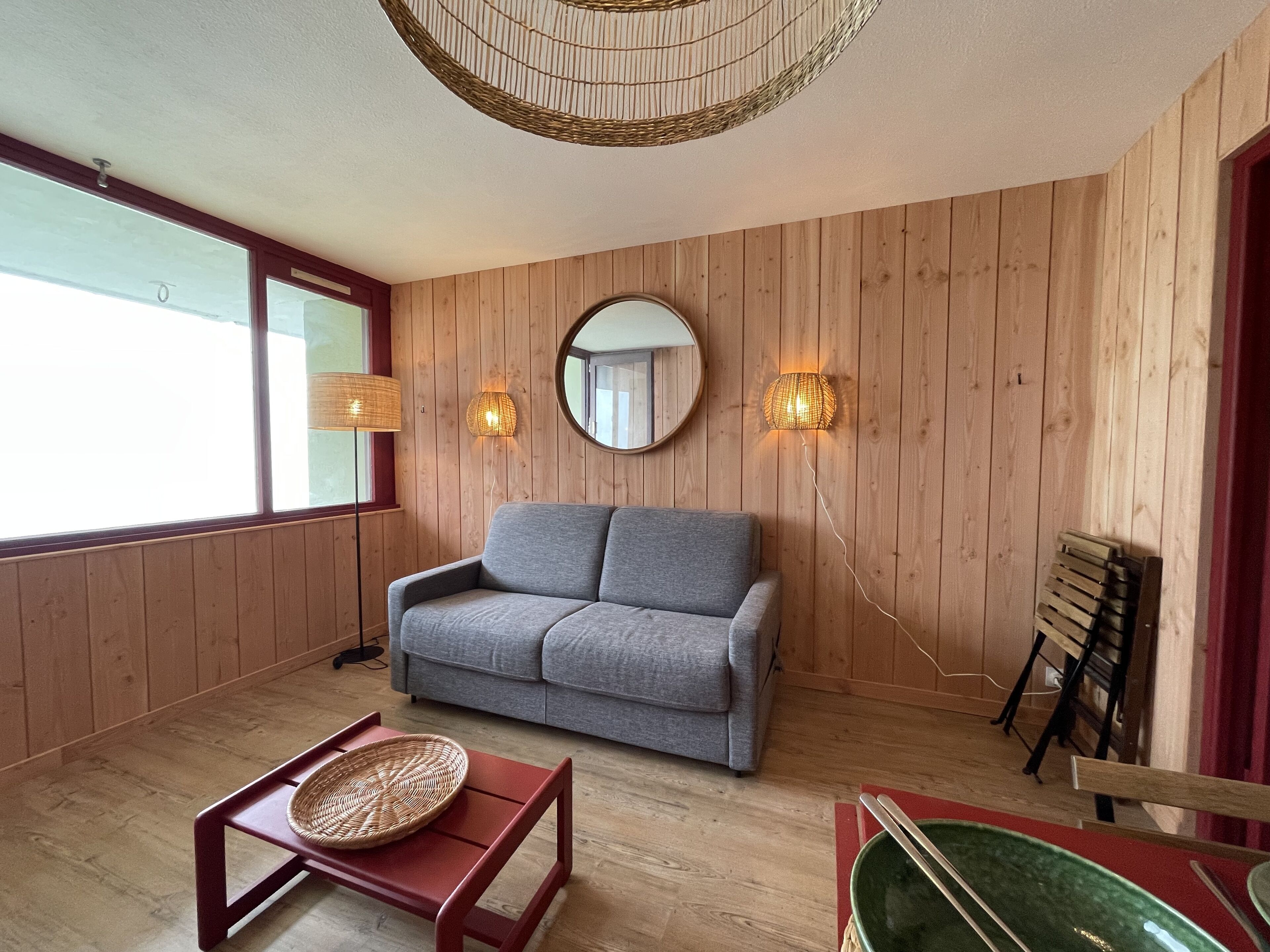 A room with a grey sofa, a wooden floor, a round mirror, and two wall-mounted lights.