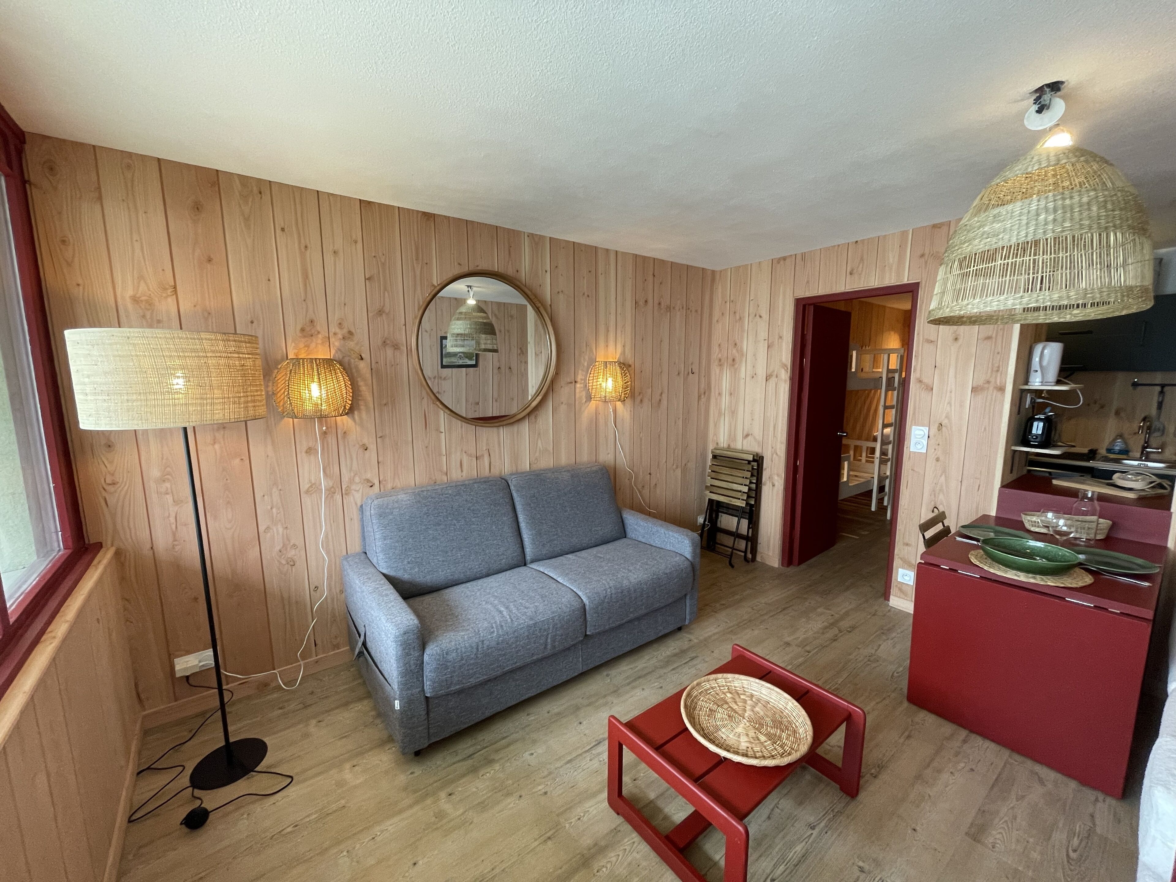 A living room with a grey sofa, a red coffee table, a wicker lamp, and a kitchen area with a red cabinet and a hanging light fixture.