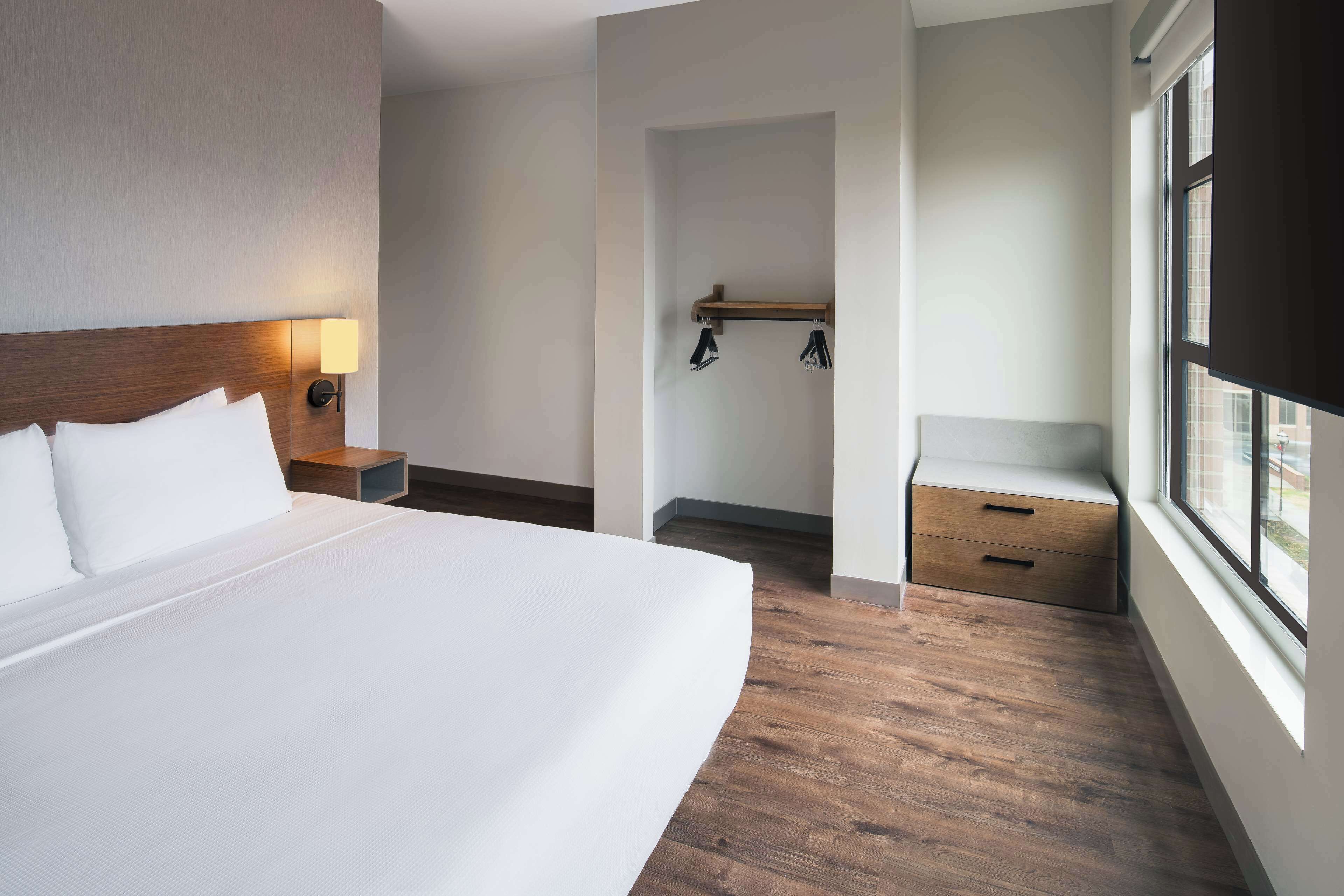 Une chambre d’hôtel avec un grand lit, une table de chevet avec une lampe, un banc en bois, un petit coin salon et une fenêtre donnant sur un paysage agréable.